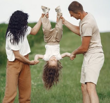 A joyful family enjoys outdoor play, lifting their child upside down in a green field.