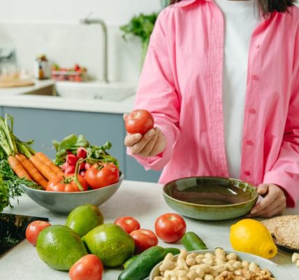 Woman in pink overshirt holds tomato in kitchen with fresh fruits and veggies.
