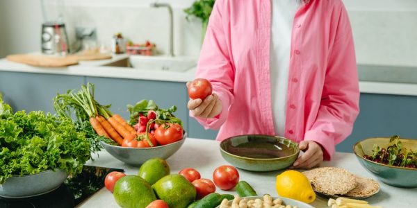 Woman in pink overshirt holds tomato in kitchen with fresh fruits and veggies.